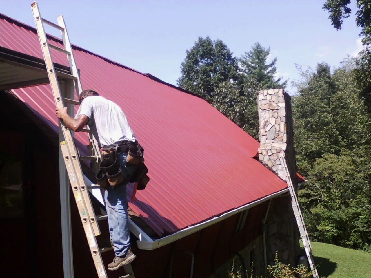 Roofer on a ladder beside a red metal roof and stone chimney — Soffit & Fascia Repair by Roofing Brothers in Echo